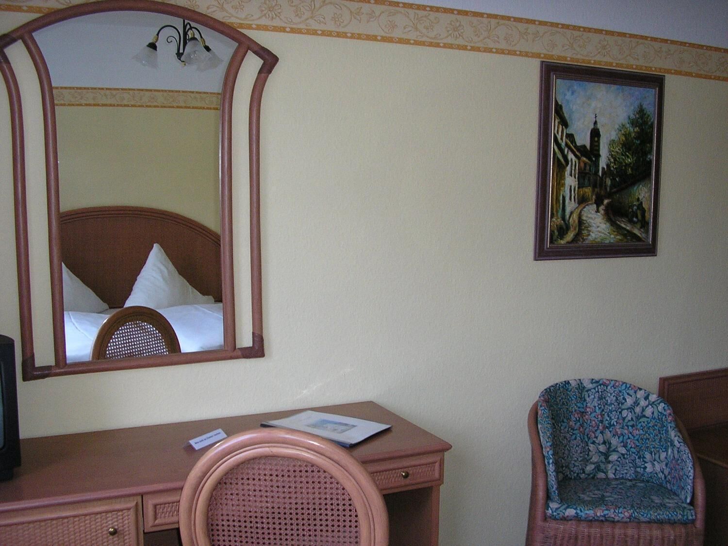 A hotel room with a bed, a desk with a chair, a mirror, and a painting on the wall.