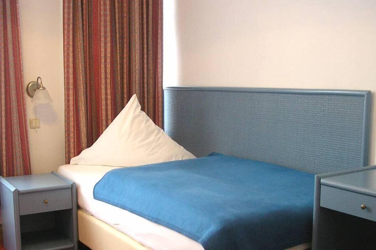 Uma cama de solteiro com cabeceira azul, travesseiro branco e cobre-leito azul. Um criado-mudo com uma luminária fica à esquerda da cama. O quarto tem uma janela com cortinas, e as paredes são de cor clara.