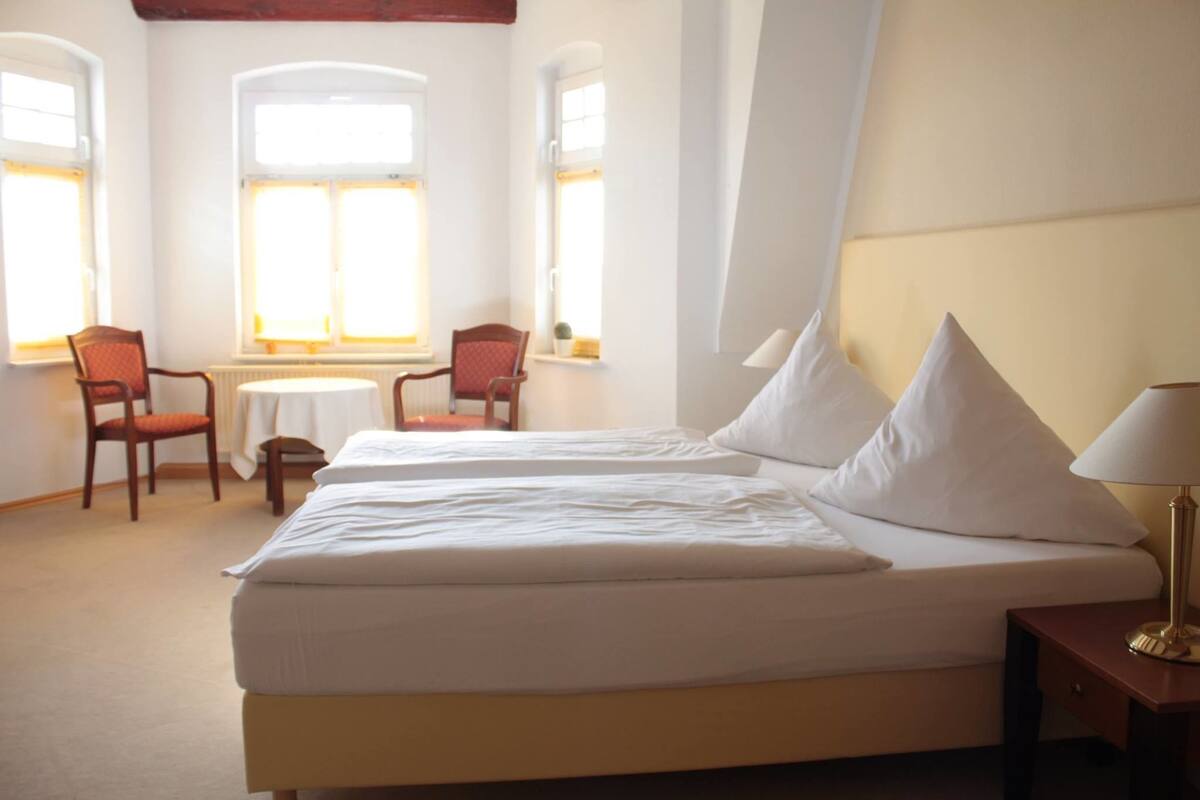 Quarto de hotel com duas camas, uma mesinha, duas cadeiras e um abajur.