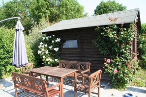 A wooden cabin with a patio table and chairs, surrounded by lush greenery and white flowers.