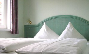 A bed with white linens and two pillows, a green headboard, and a window with purple curtains.