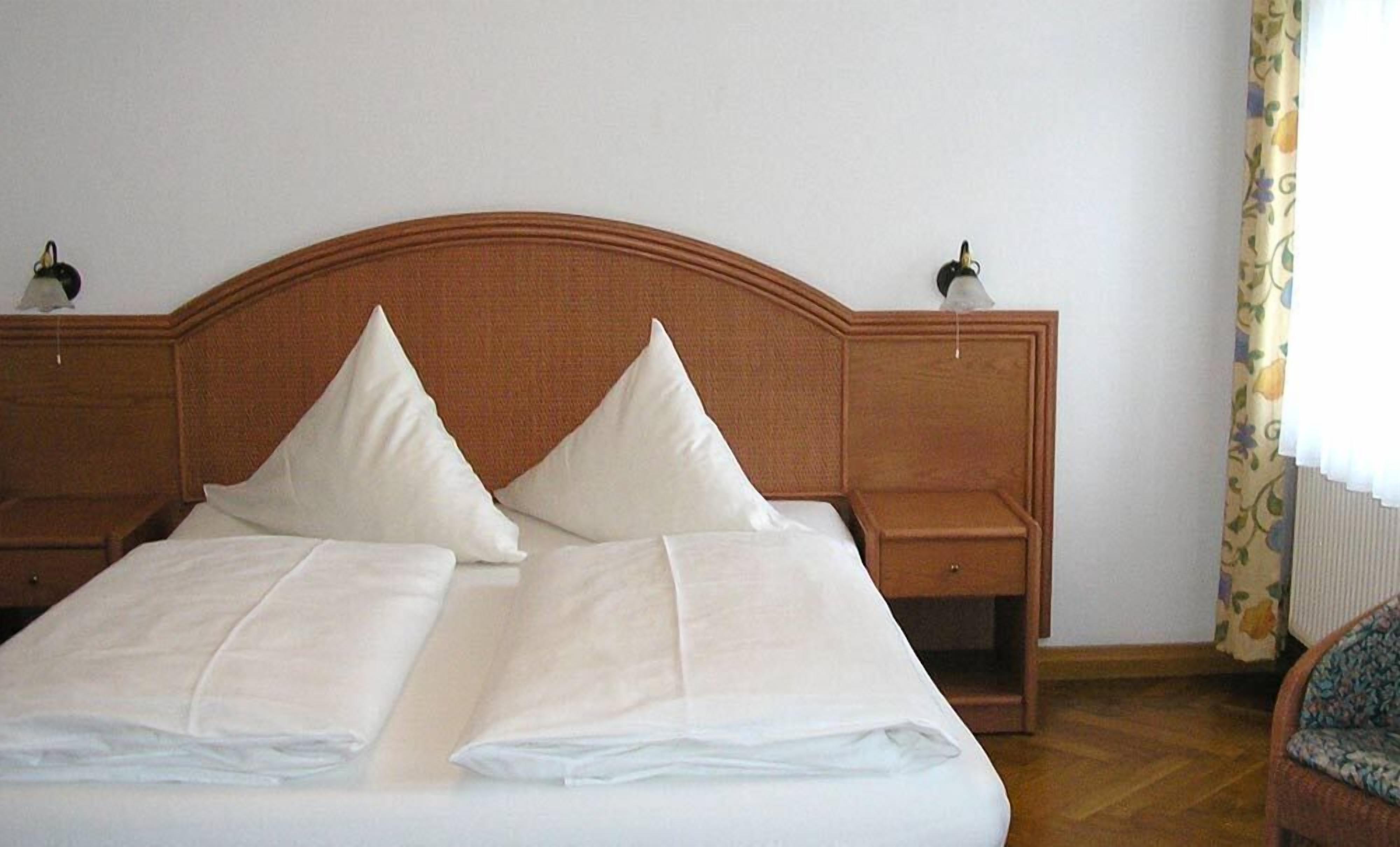 Un letto matrimoniale con lenzuola bianche e due cuscini, una testiera in legno, comodini con lampade e una tenda a motivi floreali.