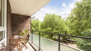 En balkon med et bord og stole, udsigt til træer, og en bygning i baggrunden.
