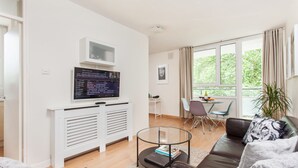 En moderne stue med et sofabord i glas, en lædersofa med pude med ulvmotiv, et ophængt tv og et spiseområde med bord og stole.