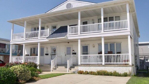 A two-story white building with a balcony on the upper floor, a covered front porch, and a small front yard. - Beach Across The Street! Great Views & Location! Sleeps 10 (Wildwood)