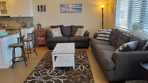 A living room with a sofa, a coffee table, and a kitchen area with a bar counter. - Beach Across The Street! Great Views & Location! Sleeps 10 (Wildwood)