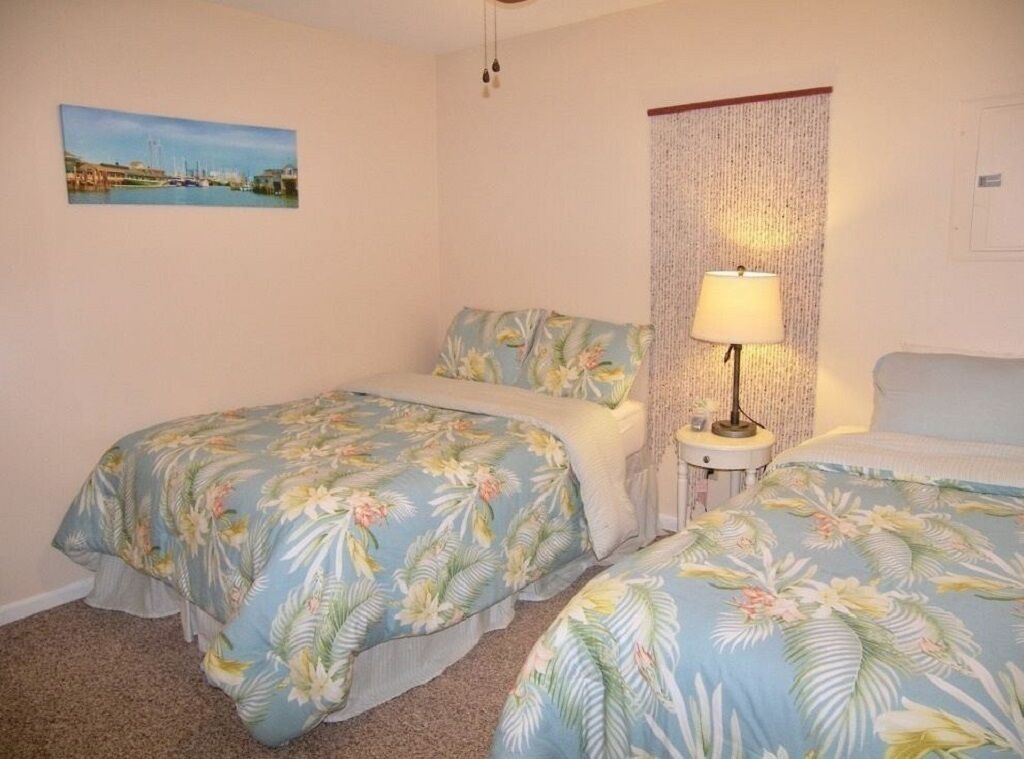 A bedroom with two beds, a nightstand with a lamp, a wall hanging, and a picture of a coastal scene.