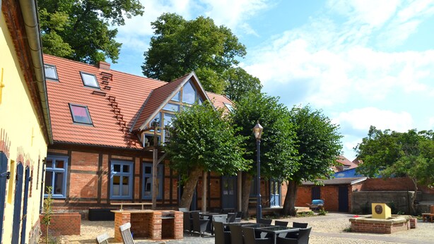 A courtyard with outdoor seating, a brick building with a sloped roof, and trees. - Kulturhof (Lenzen)