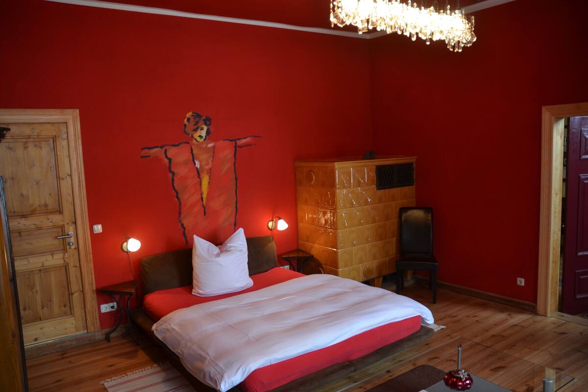 Une chambre avec un mur rouge, un lit en bois, une table de chevet, une commode et un lustre.