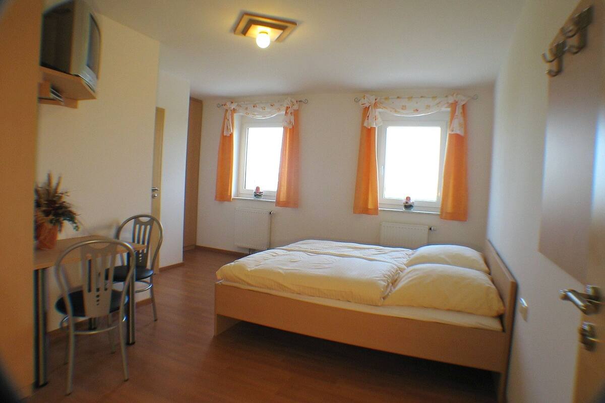 A bedroom with a single bed, two windows with curtains, a radiator, a TV mounted on the wall, and a chair.