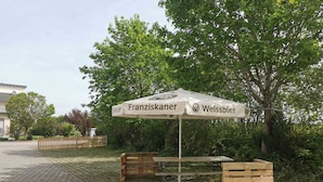 Un espace extérieur aménagé avec une table et des bancs, un parasol arborant le logo de la marque « Franziskaner Weissbier », et un chemin pavé.
