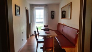 Un couloir avec un plancher en bois, une table à manger avec des chaises, un canapé rouge, un radiateur, une horloge et une fenêtre avec des rideaux.