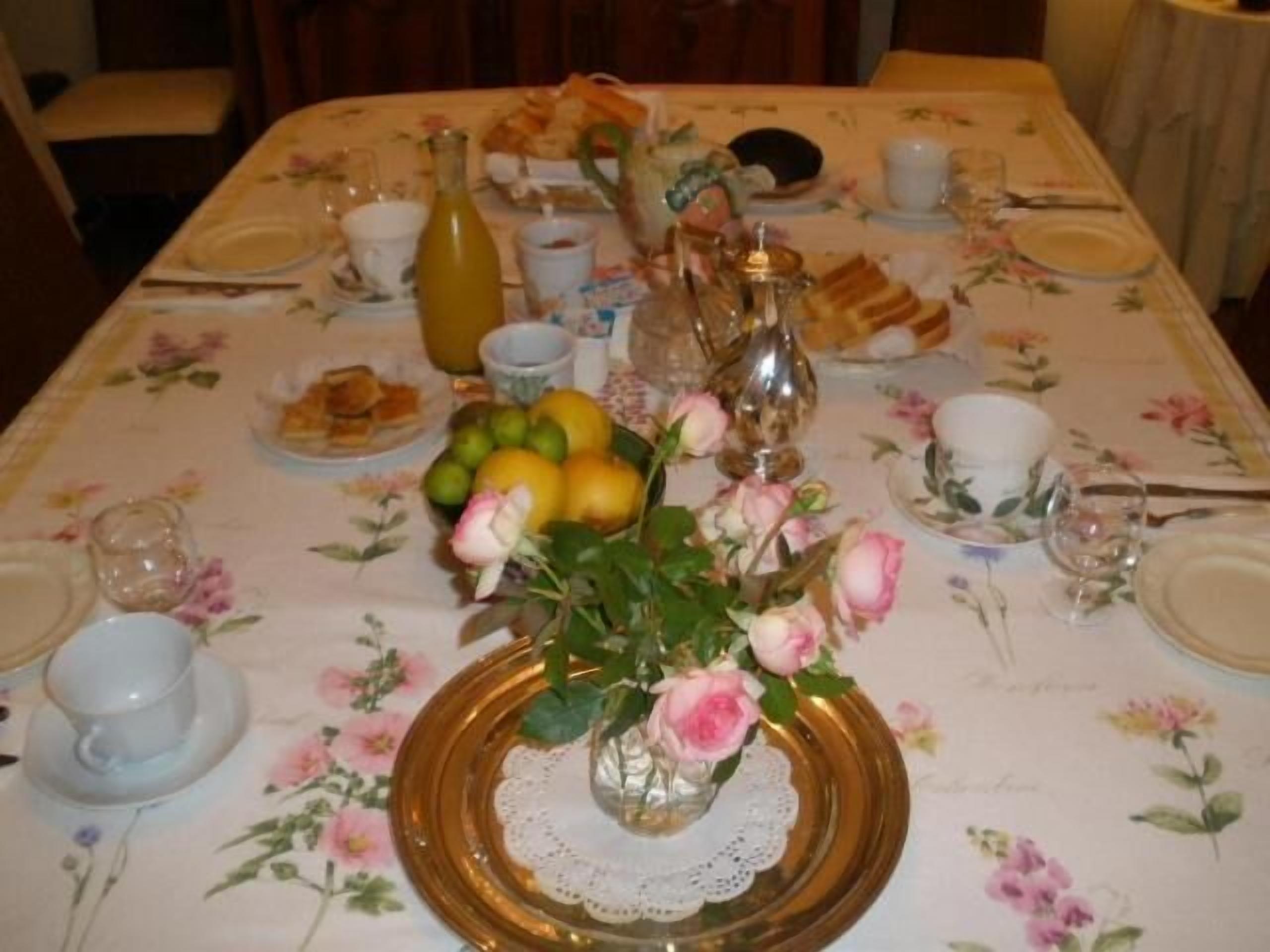 Ein Esstisch, gedeckt mit einer blumigen Tischdecke, Tellern, Gläsern, einer Rosenvase und einer Flasche.