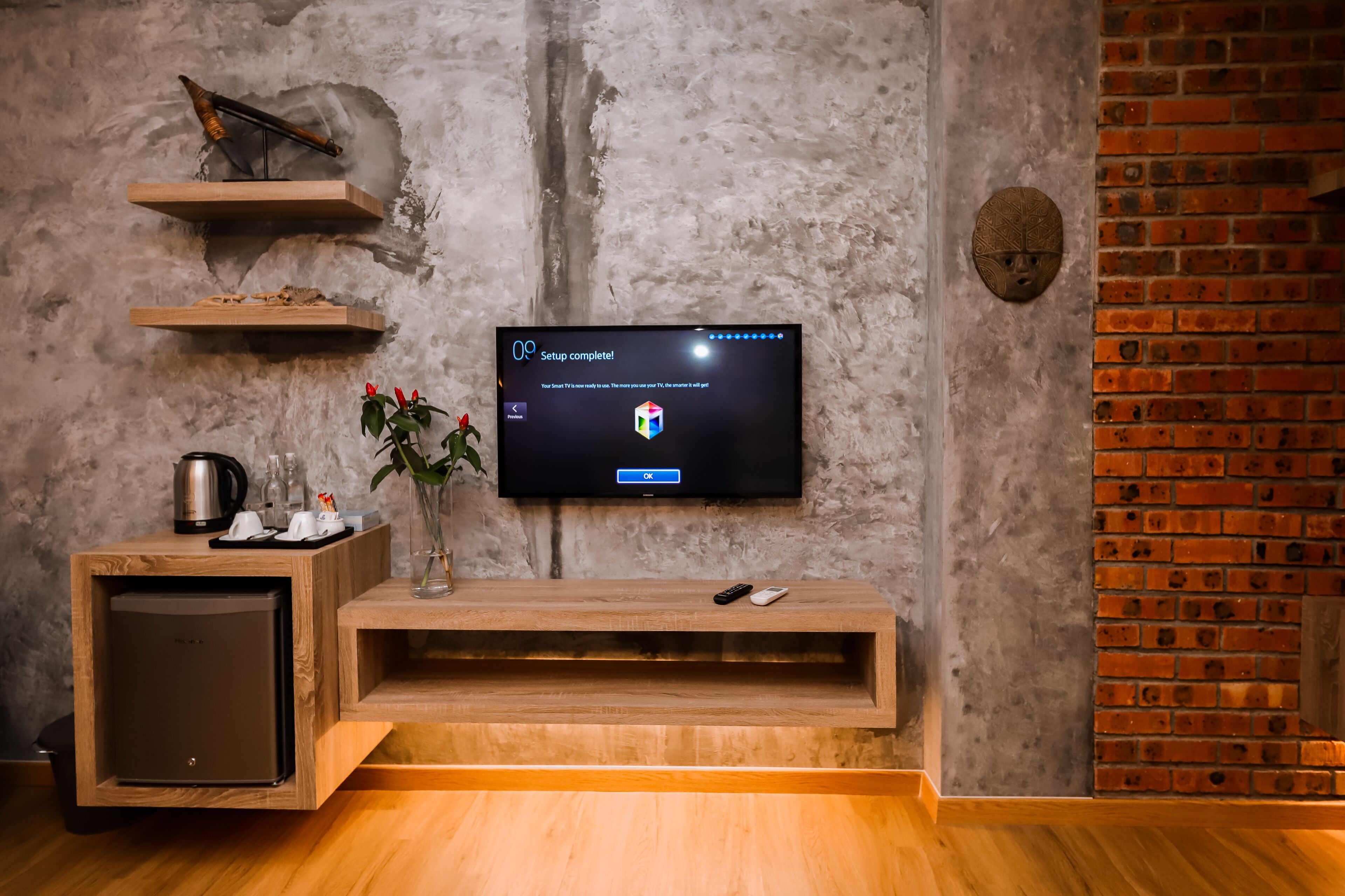Ein modernes Wohnzimmer mit Flachbildfernseher an einer strukturierten Wand, Holzmöbeln, einer Kaffeemaschine und einem kleinen Kühlschrank.