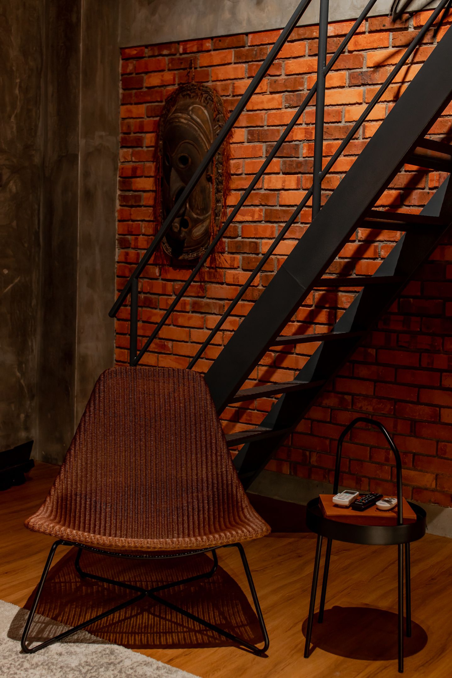 A staircase with a brick wall and a wooden floor, a woven chair, and a small table with a phone and remote control.