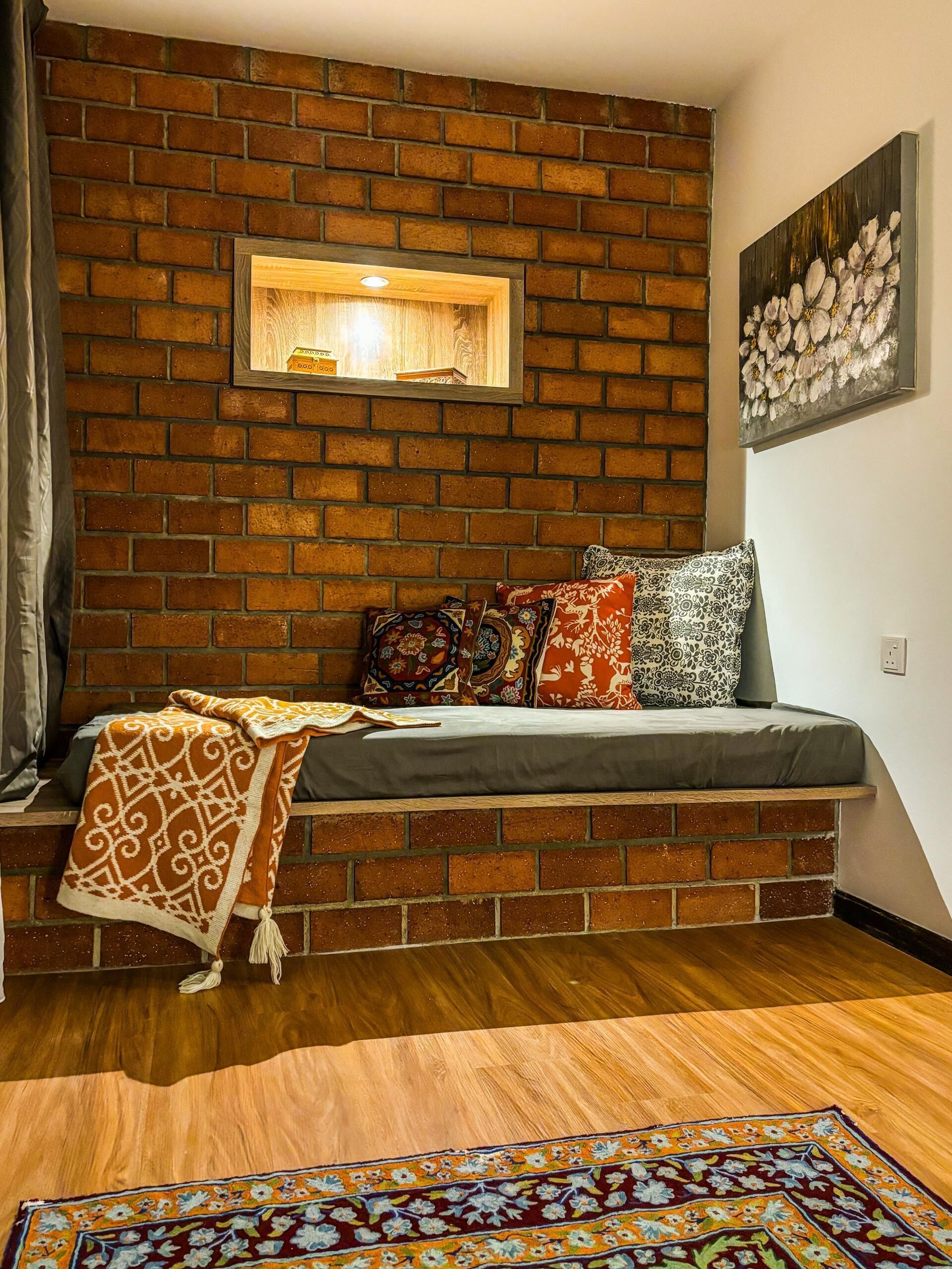 Ein gemütliches Zimmer mit Backsteinwand, einer eingebauten Sitzbank mit Kissen, einem gemusterten Teppich und einem gerahmten Bild an der Wand.