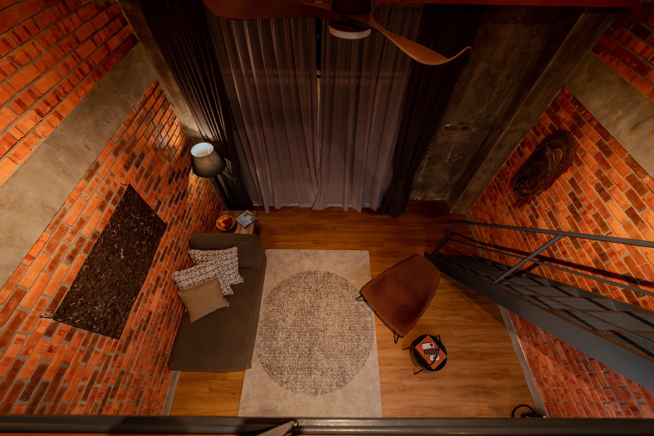 Habitación con pared de ladrillo, suelo de madera, alfombra circular, sofá marrón, sillón marrón, pequeña mesa con lámpara y ventilador de techo.
