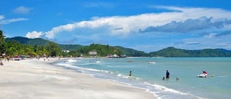 A beach with clear waters, people swimming, and a few boats.