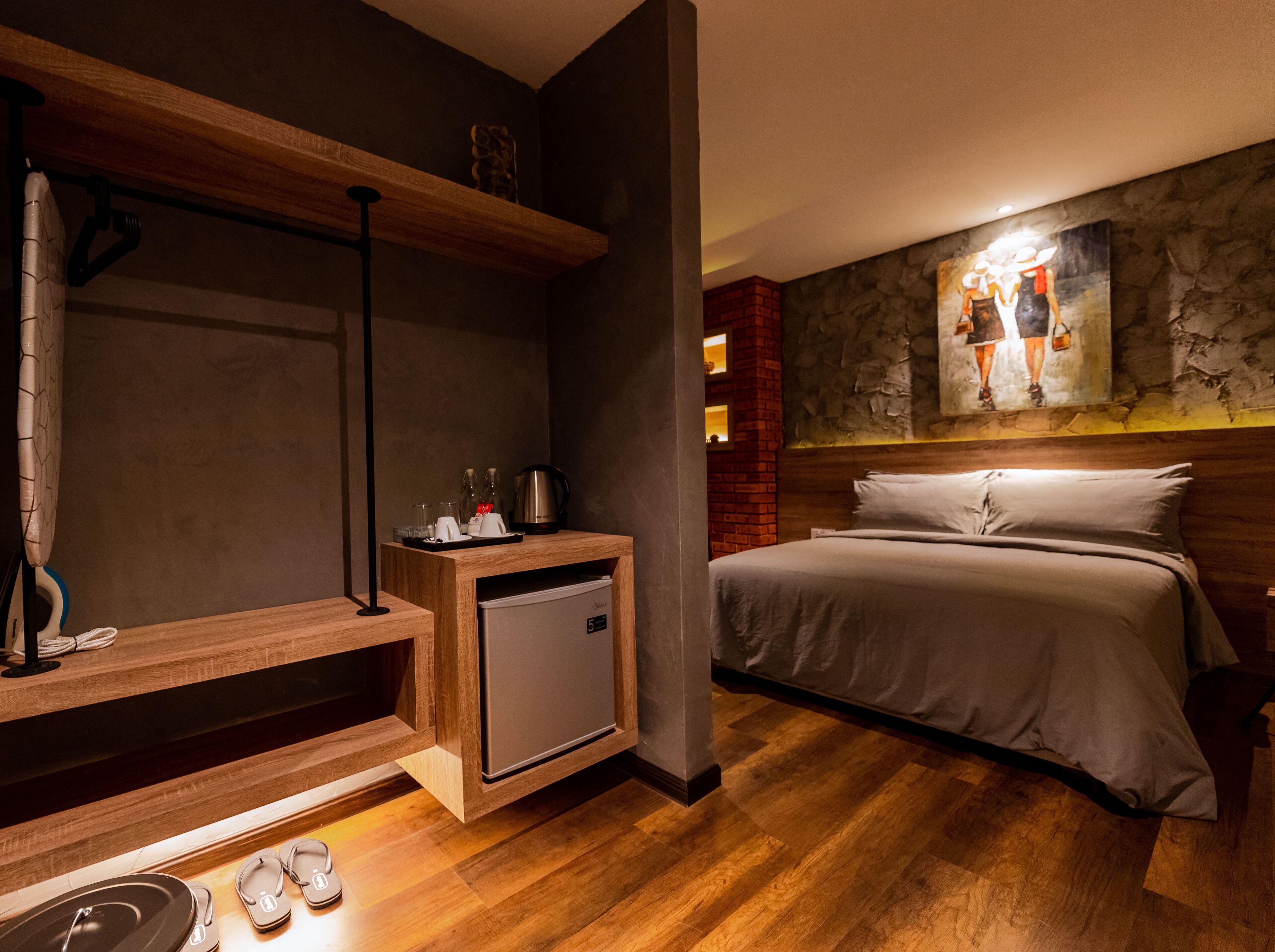 A modern hotel room with a bed, a small kitchenette, a wooden shelf, and a stone wall with a painting.