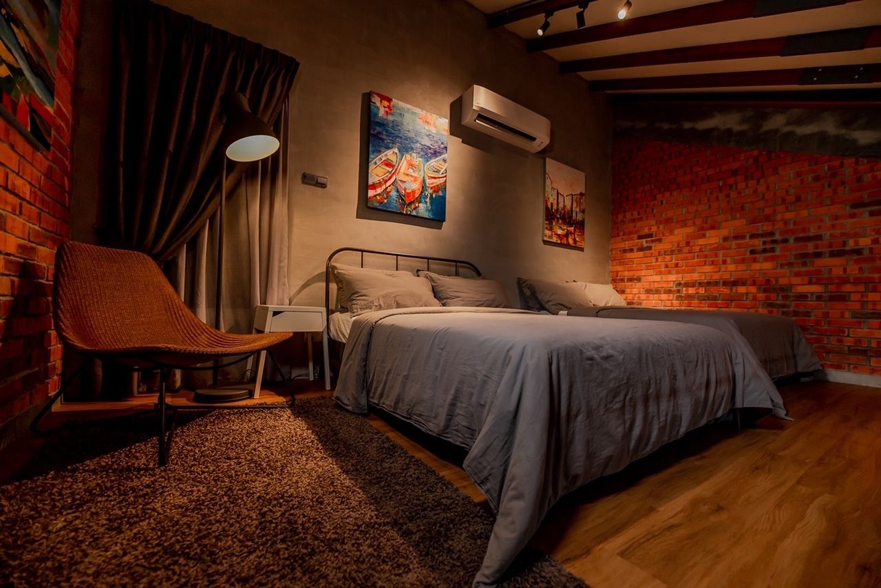 A bedroom with a brick wall, a bed with grey bedding, a chair, and artwork on the wall.