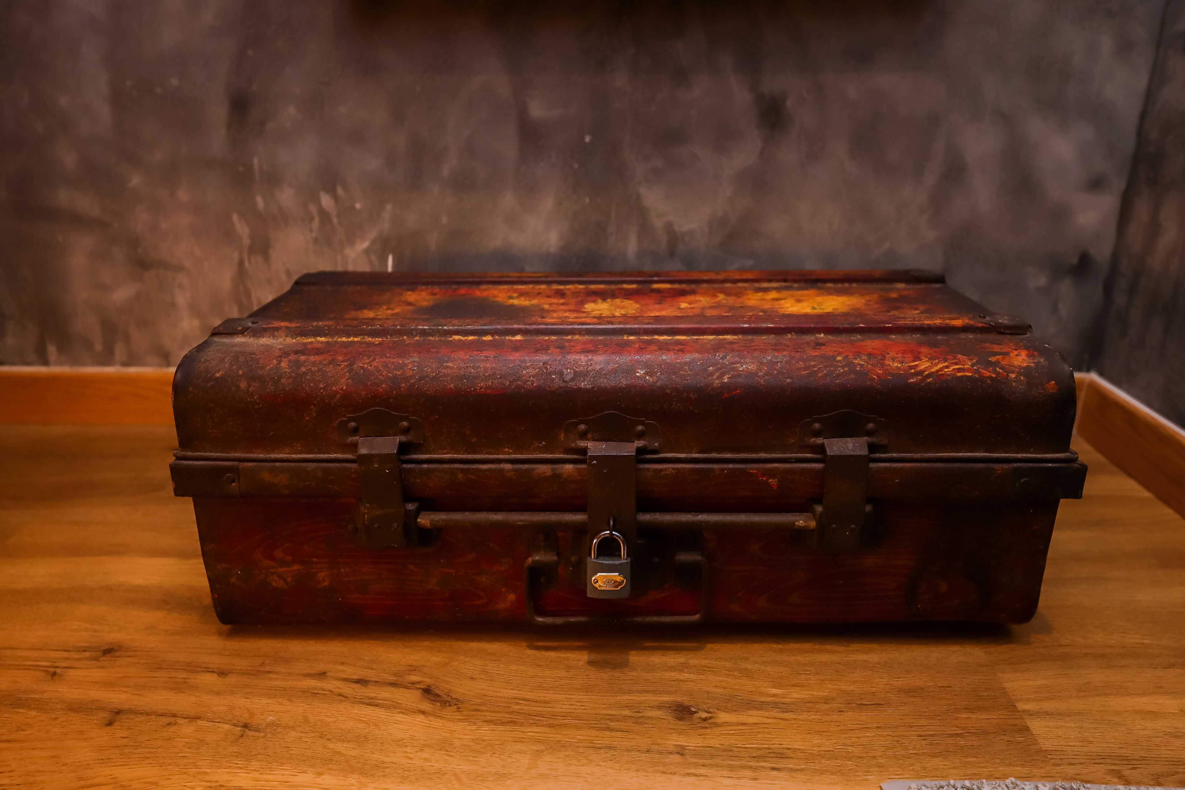 A vintage, metal suitcase with a lock on a wooden floor against a dark wall.
