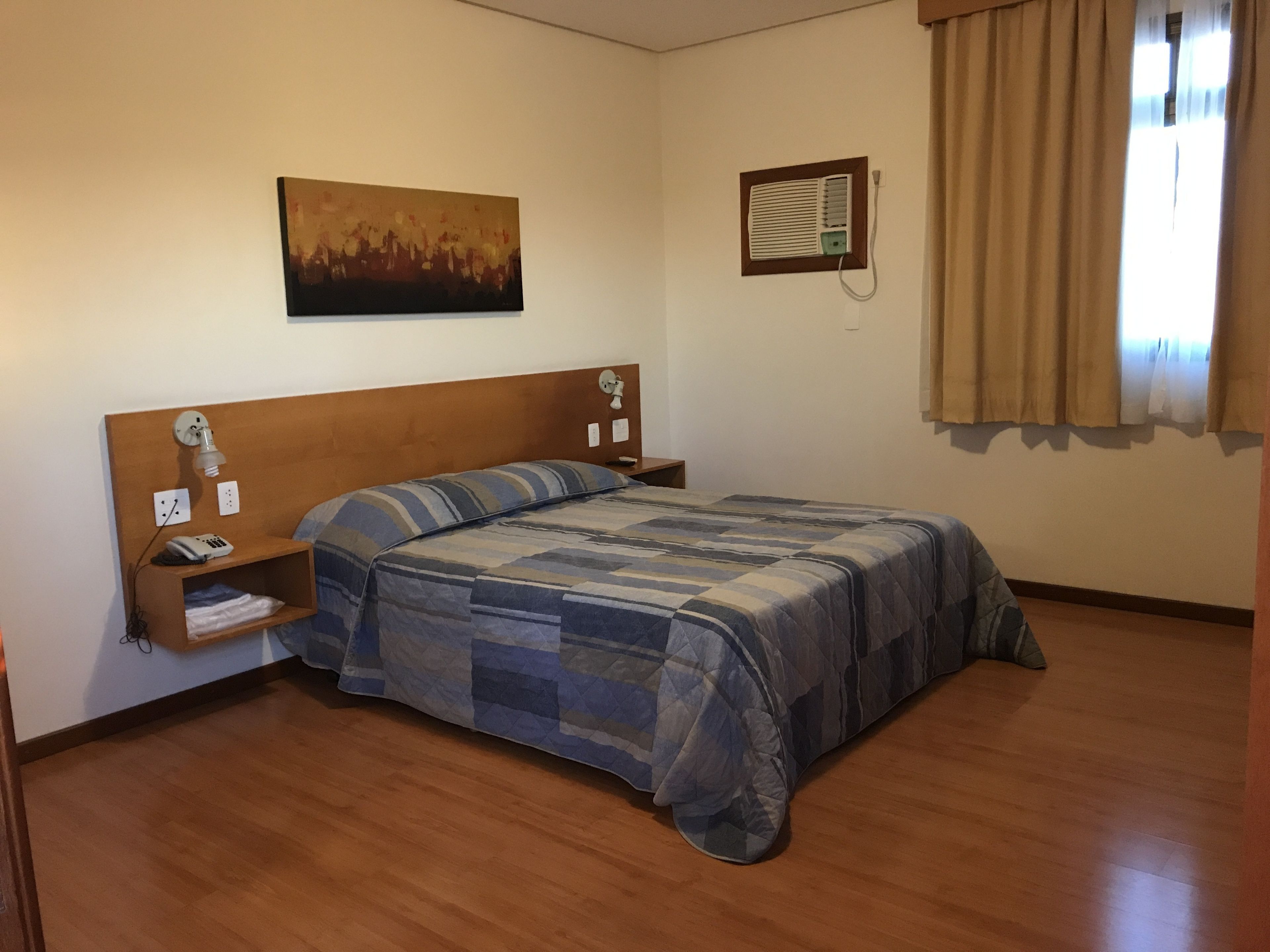 A bedroom with a wooden headboard, a bed with a striped blanket, a bedside table with a lamp, a wall-mounted air conditioner, and a painting on the wall.