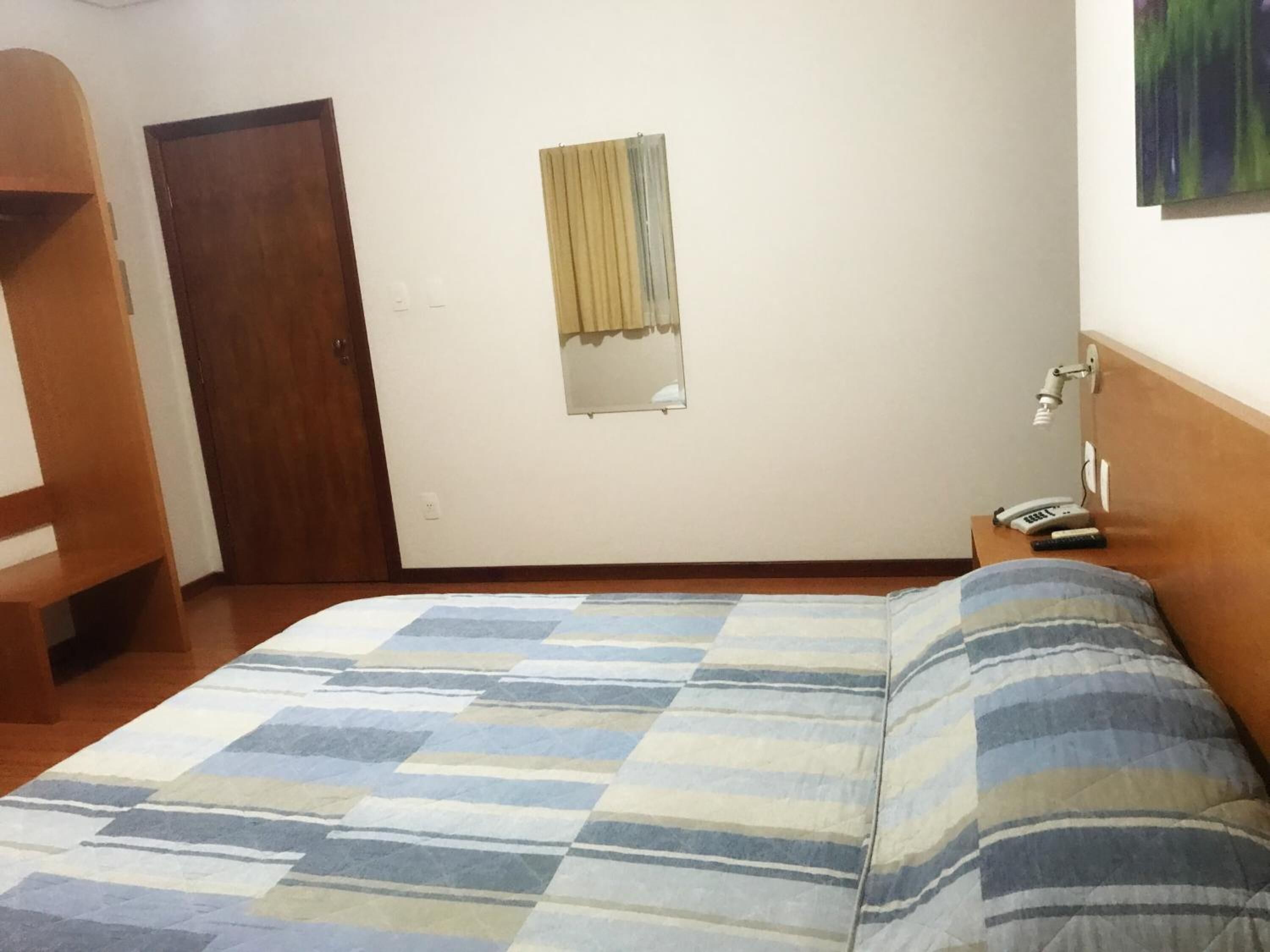 Ein Schlafzimmer mit einem Bett, einer Holztür, einem kleinen Fenster mit Vorhang und einem Nachttisch mit Lampe und Telefon.
