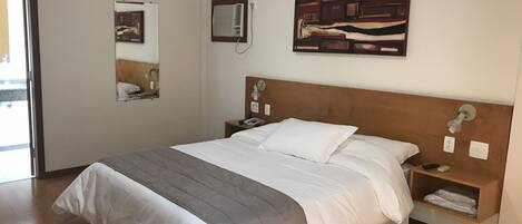 Una cama bien hecha con edredón gris y blanco, cabecera de madera, mesitas de noche con lámparas, aire acondicionado de pared y un cuadro enmarcado sobre la cama.