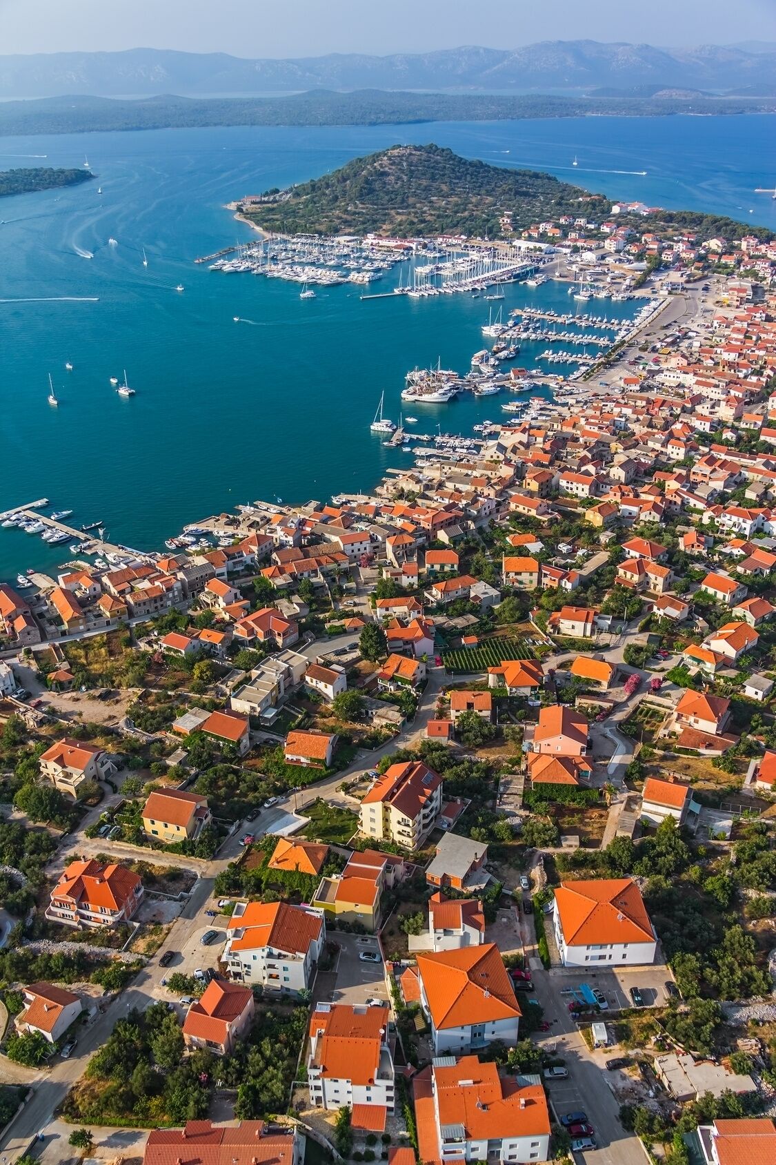 An aerial view of a coastal town with a marina, houses with red roofs, and a hilly island in the background.
