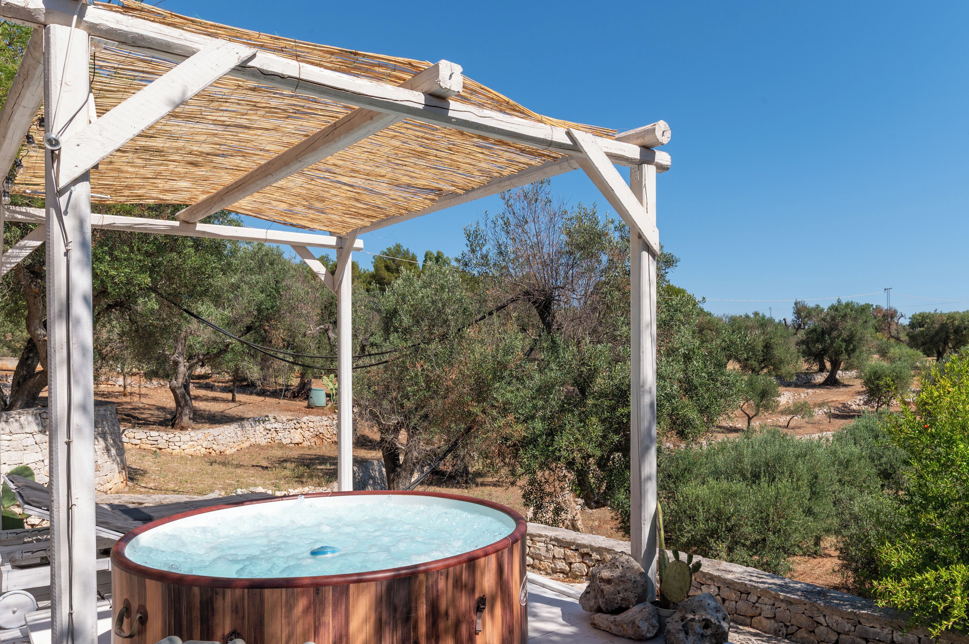 Una vasca idromassaggio in legno sotto una pergola, con vista sulle olive e un muro in pietra.