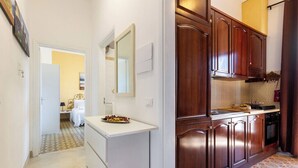 A hallway leading to a bedroom with a bed, a small table with fruit, and a mirror. The room has a white dresser and a wooden cabinet with a stove. - Appartamento La Rosa Dei Venti (Ostuni)