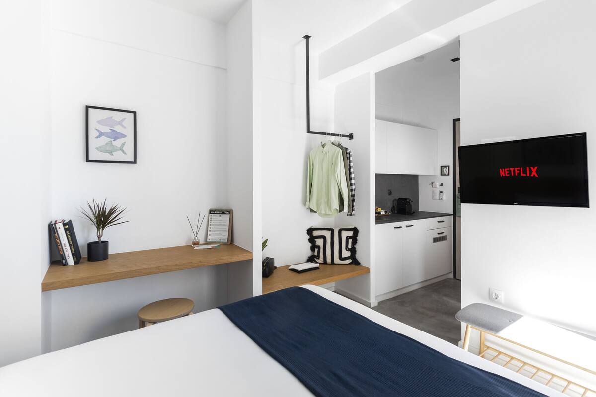 A modern hotel room with a bed, a desk with a plant, a wall-mounted TV, and a view into the kitchen.