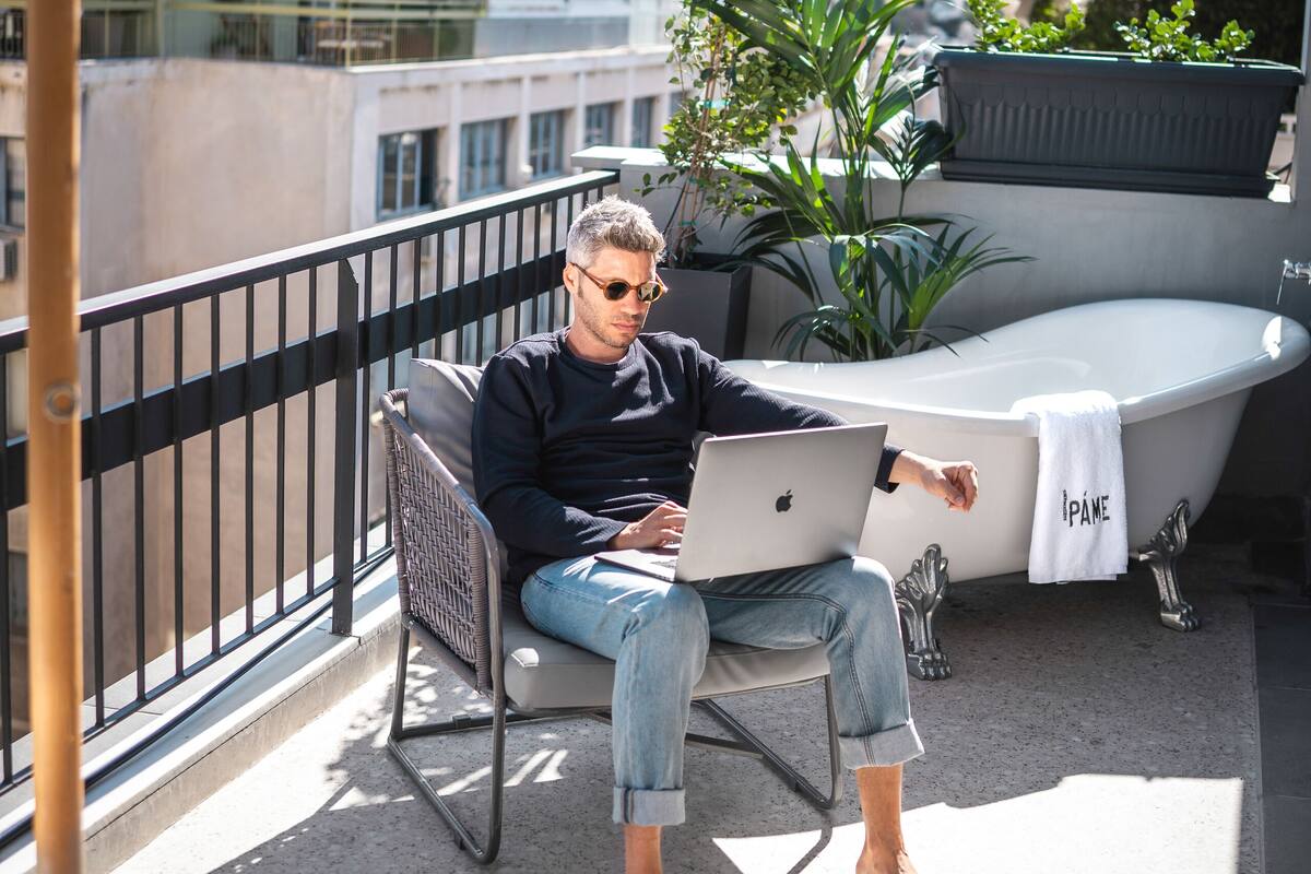 A person sitting on a balcony chair with a laptop, a bathtub, and potted plants.