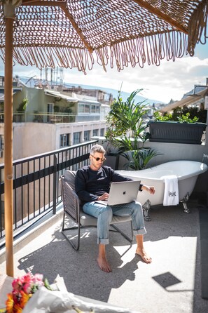 A person sitting on a balcony chair with a laptop, a bathtub, and potted plants. - Pame House (Athens)