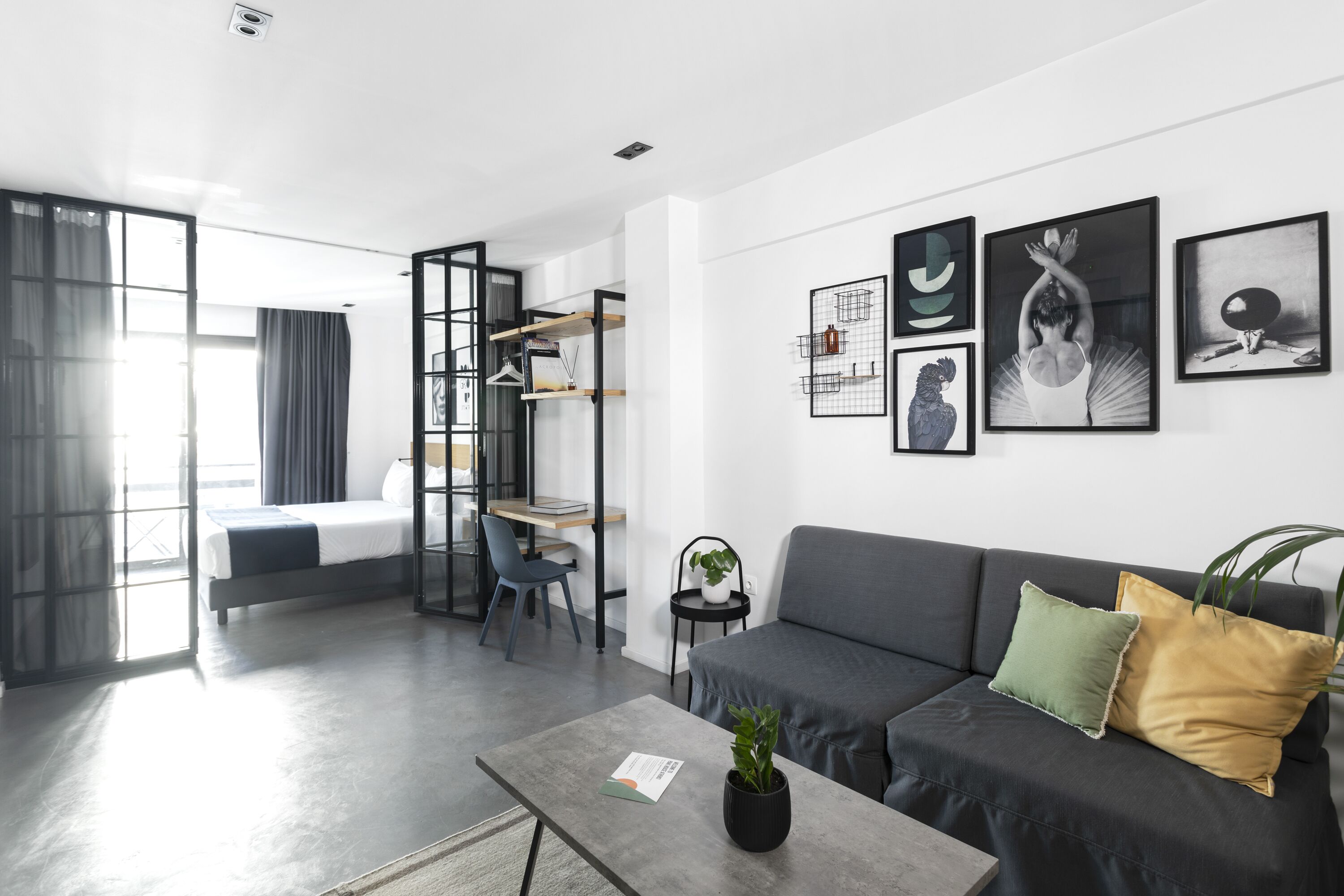 A modern living room with a grey sofa, a coffee table with a plant, and a desk with a chair. There are framed pictures on the wall and a large window with curtains.