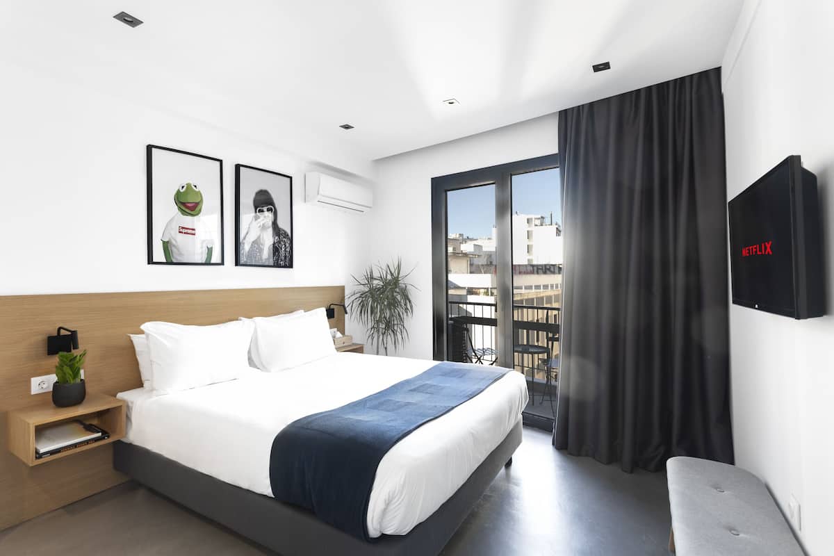 A modern hotel room with a large bed, a bedside table with a plant, a wall-mounted TV, and a balcony with a view of the city.