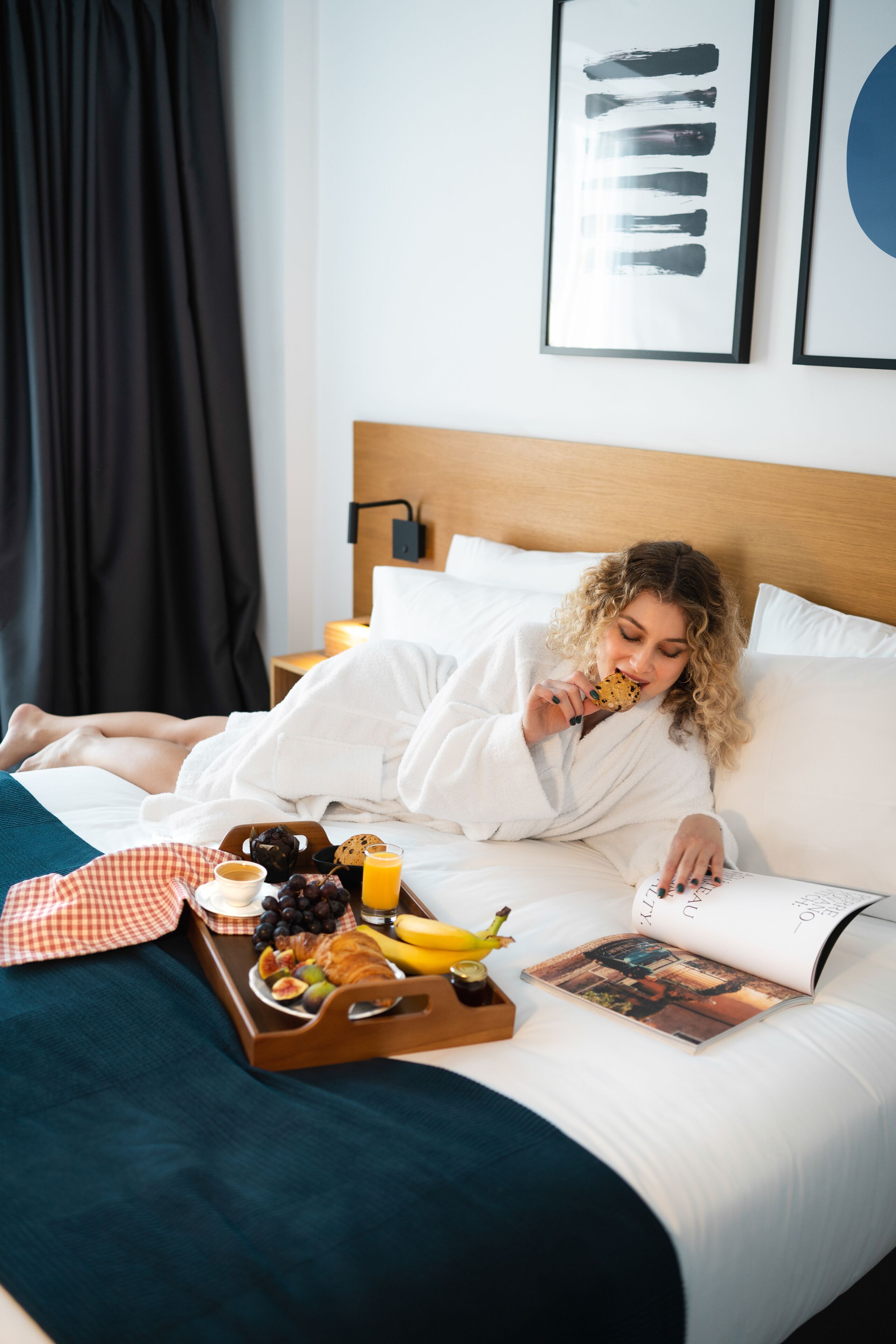 A person lying in bed with a tray of breakfast items, a book, and a pillow.