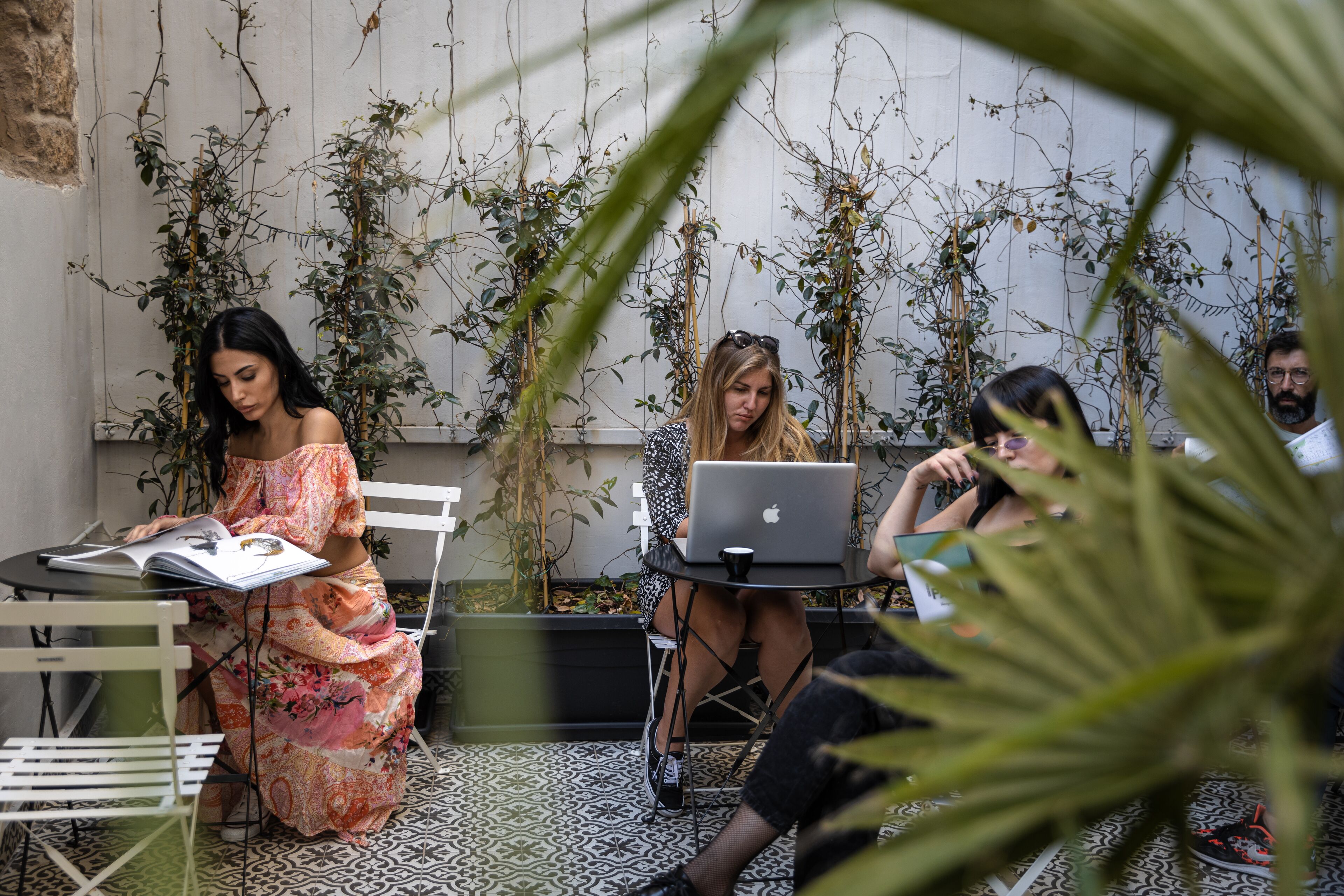 A group of people working at tables with laptops in an outdoor setting with plants and a tiled floor.