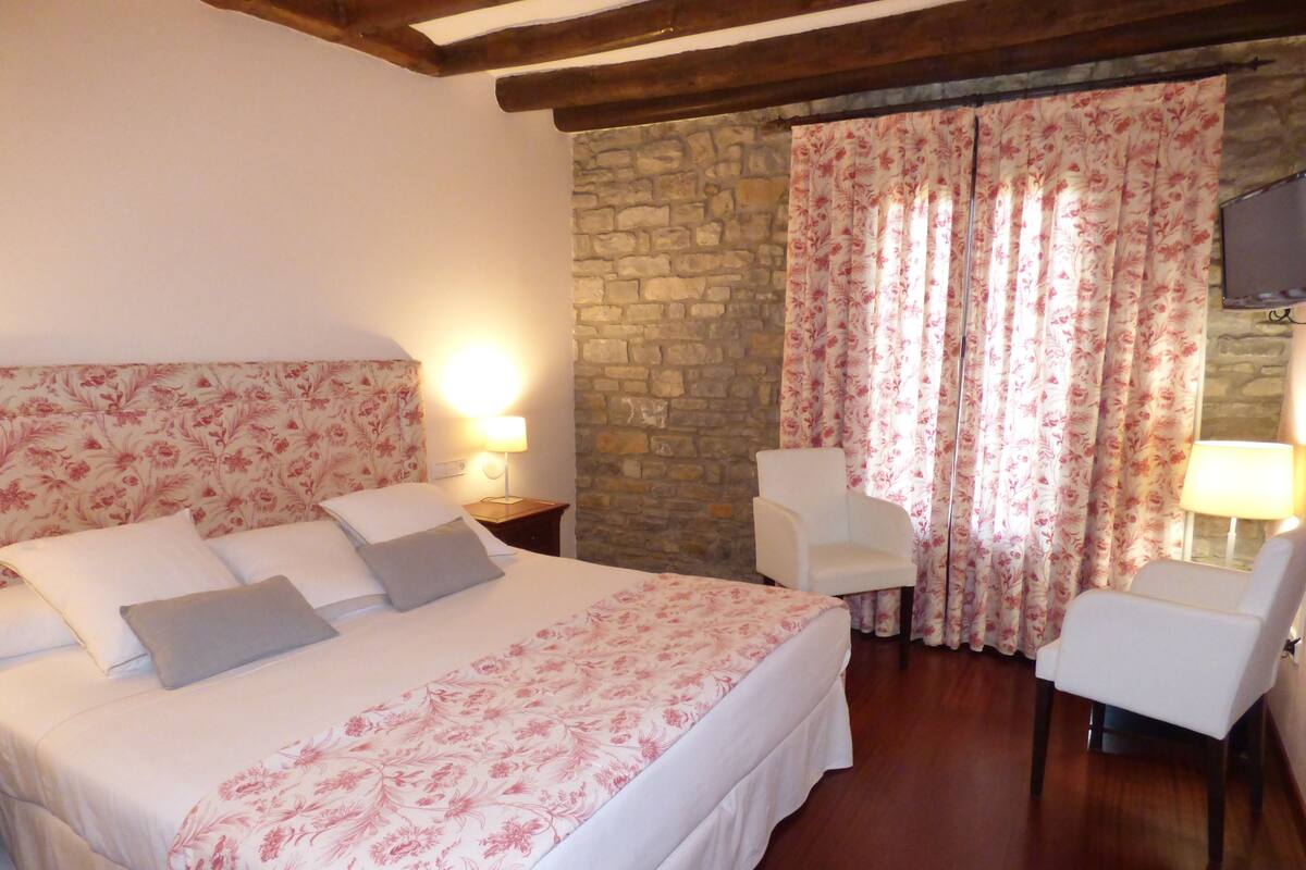 A bedroom with a large bed, two bedside tables with lamps, a stone wall, and a chair by the window with floral curtains.