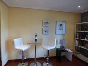 A room with two white bar stools, a glass table with a bottle and glasses, framed artwork on the wall, and a bookshelf filled with books. - Hotel Los Arcos (Ainsa)