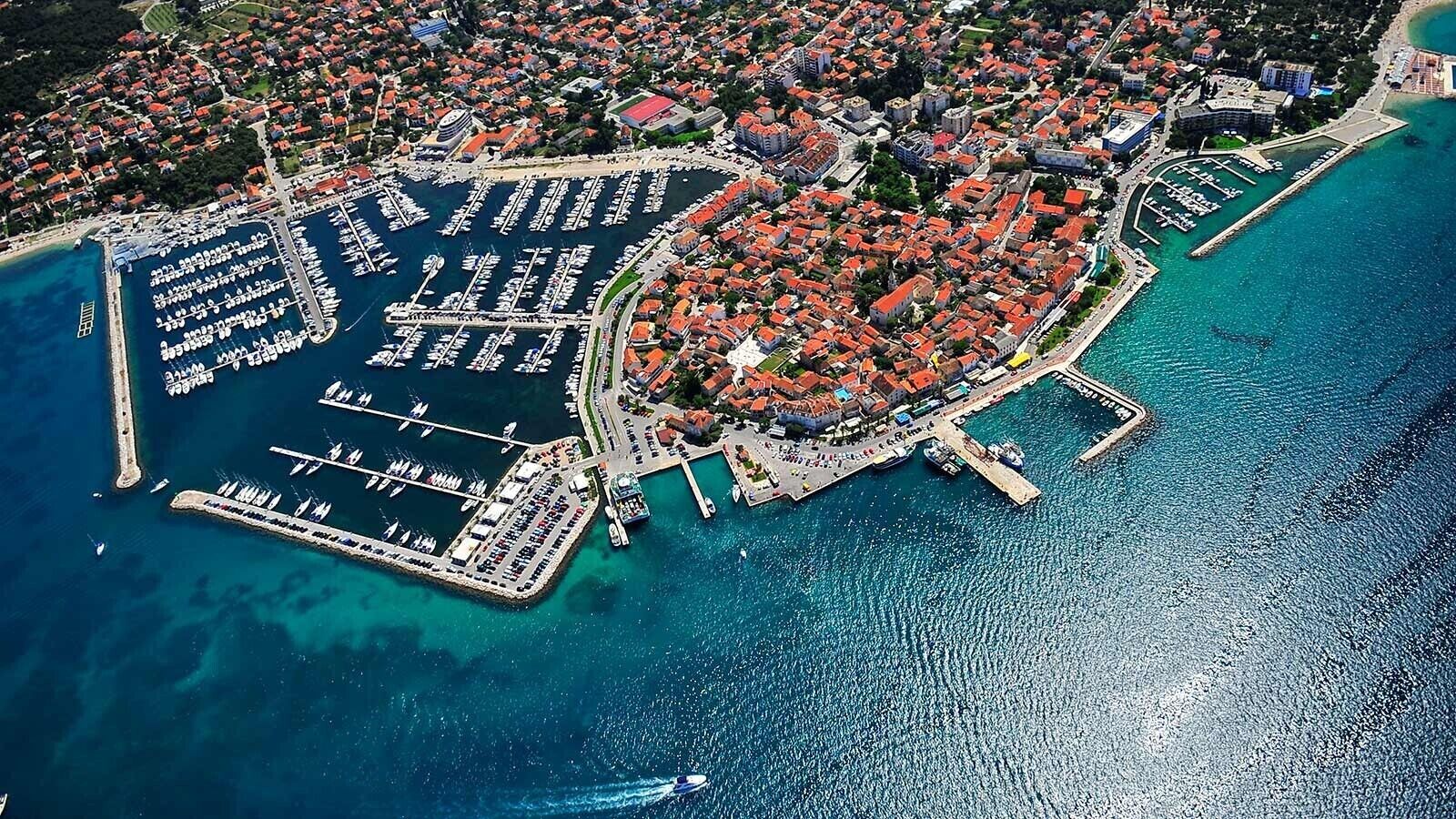Luchtbeeld van een kustplaatsje met een jachthaven, woonwijken en een klein schiereiland met gebouwen en steigers.