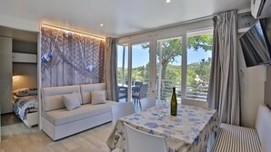 Un salon moderne comprenant un canapé, une table à manger et des chaises, de grandes fenêtres donnant sur un paysage verdoyant, et un téléviseur fixé au mur.