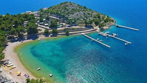 Vue aérienne d’une station balnéaire avec une baie aux eaux turquoise et limpides, une jetée où sont amarrés des bateaux, et une petite île parsemée de bâtiments et de végétation.