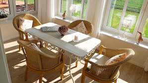 Un coin repas avec une table blanche et des chaises en osier, de grandes fenêtres donnant sur une pelouse verdoyante et un lustre.