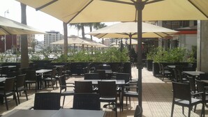 Un café en plein air avec des tables et des chaises placées sous de grands parasols, entouré de verdure et avec des bâtiments en arrière-plan.