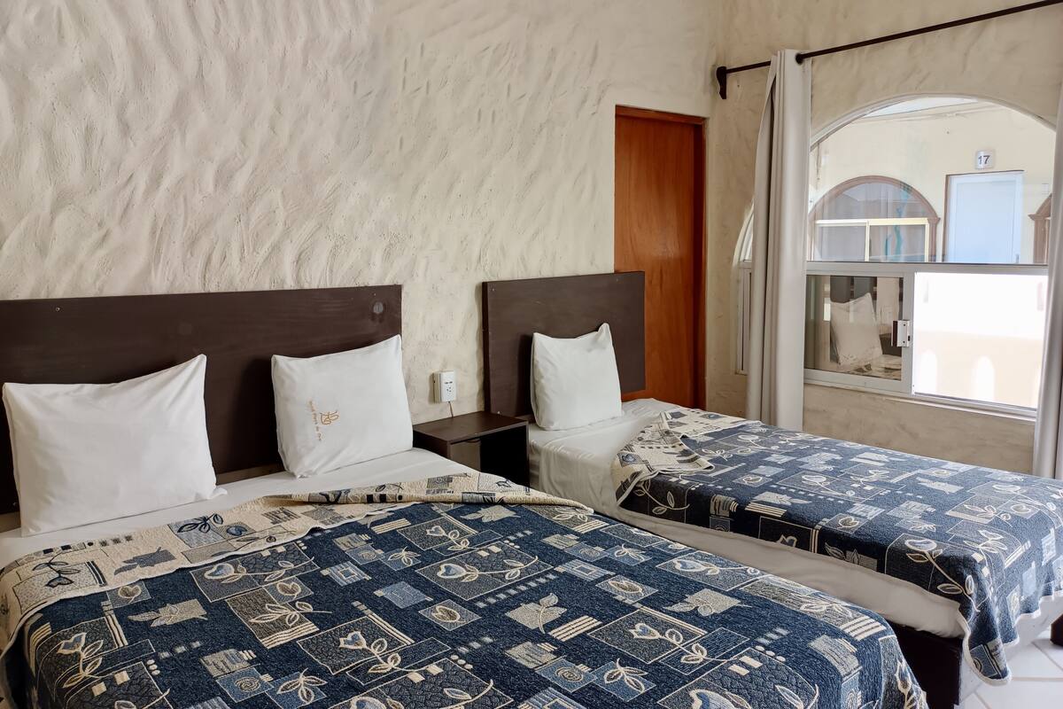 Habitación de hotel con dos camas, cada una con una colcha estampada, cabeceros de madera y almohadas blancas. Se aprecia una ventana con vistas al exterior.