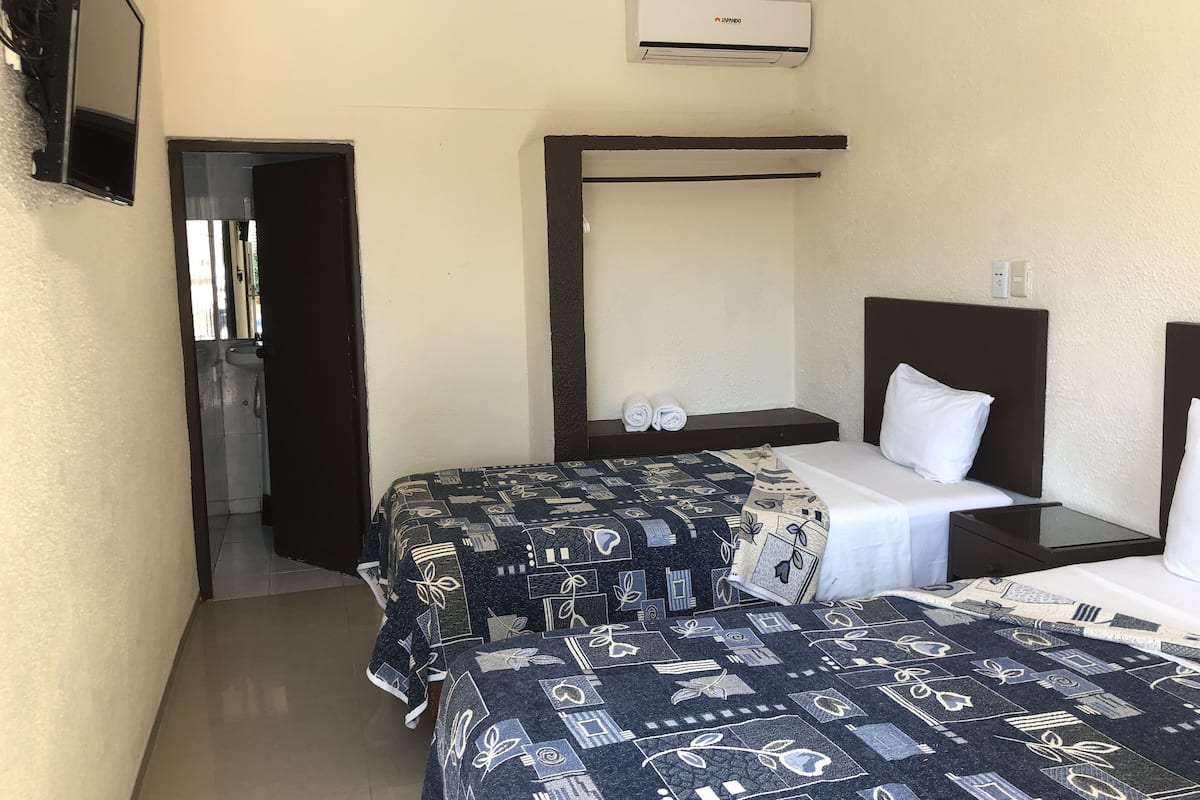 Una habitación de hotel compacta con dos camas, una mesita de noche pequeña, un aire acondicionado de pared y un baño visible a través de una puerta abierta.