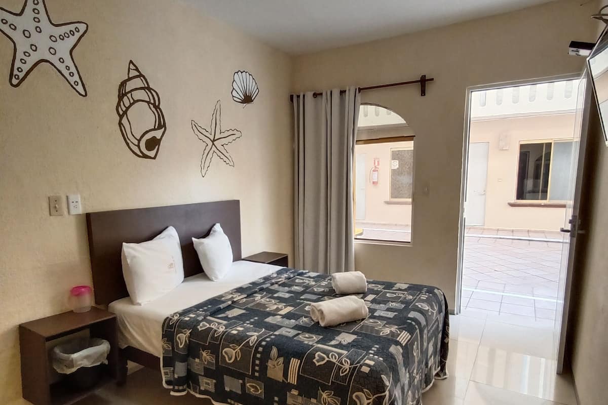 Habitación de hotel con cama, mesita de noche, pared decorada con estrellas de mar y conchas, ventana con cortinas y vista al exterior.