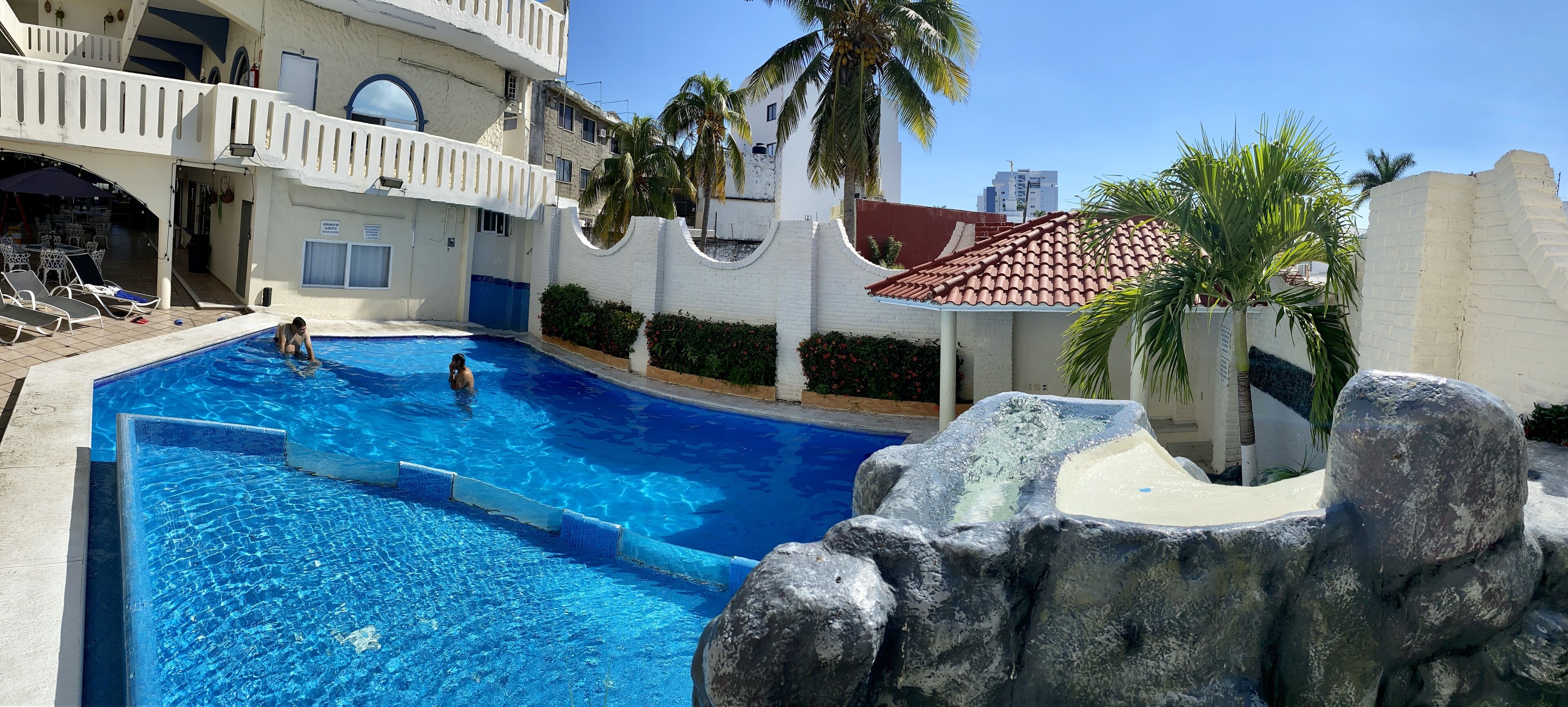 Una piscina con una formación rocosa, palmeras y un edificio con balcones.
