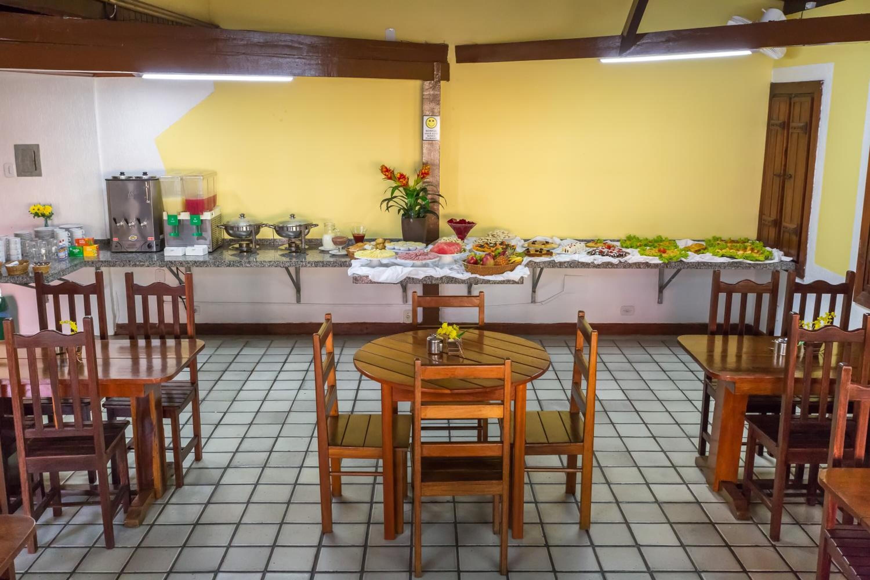 A dining area with a round wooden table and chairs, a tiled floor, a yellow wall, and a counter with food and drinks.